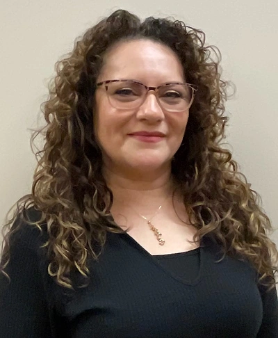 Woman with curly brown hair and glasses smiling at camera in dental office setting.