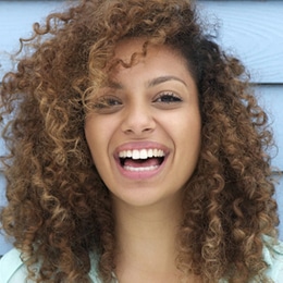 Smiling woman with curly brown hair against light blue background for dental practice.
