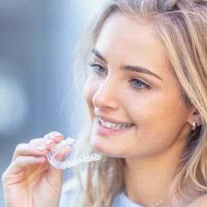 Woman smiling while holding clear dental aligners against blurred background.