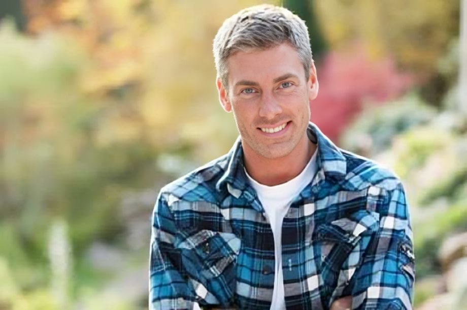 Middle-aged man in blue plaid shirt smiling outdoors with blurred garden background.