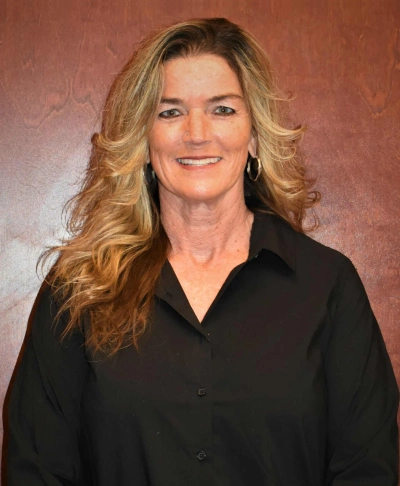 Professional headshot of a woman with blonde wavy hair wearing a black collared shirt against a warm brown background.