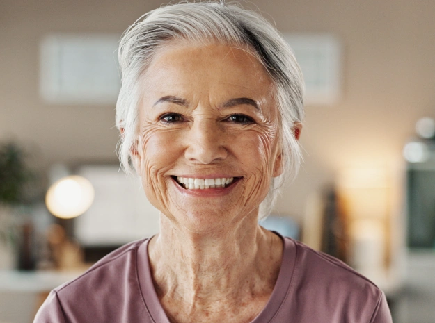 Smiling senior woman with gray hair in dental office setting wearing purple shirt.