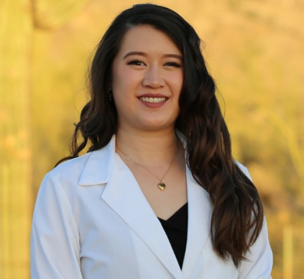 Female dental professional smiling at camera wearing white coat against yellow background.