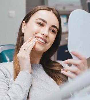 Woman smiling while examining teeth in mirror at dental office.