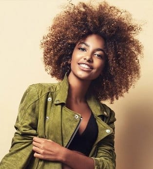 Woman with curly brown hair wearing olive green jacket, smiling against neutral beige background.