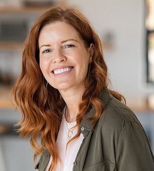 Smiling woman with reddish-brown hair in olive jacket at dental office.