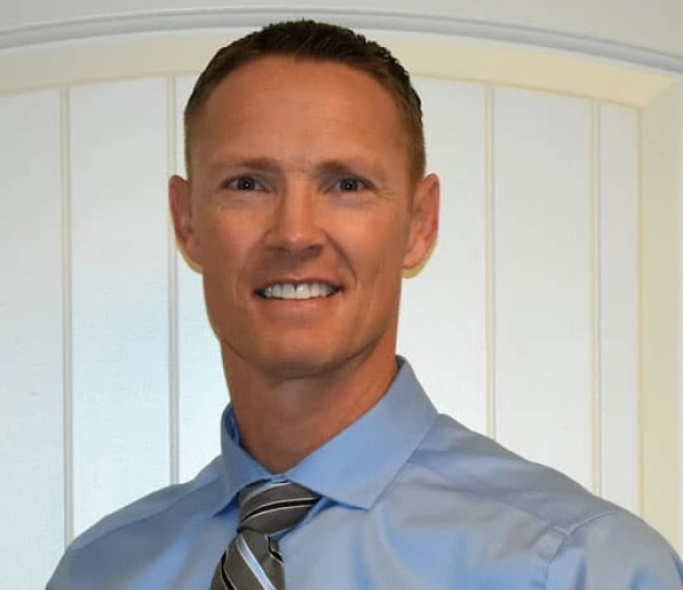 Professional headshot of smiling man in blue shirt and tie in dental office setting.