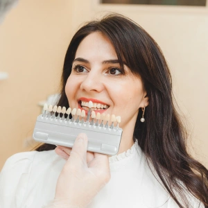 Woman holding dental shade guide against teeth, smiling in dental office setting.