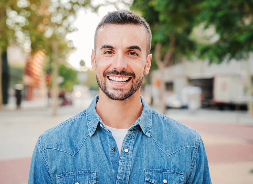 Smiling man in denim shirt standing outdoors on brick pathway in front of blurred urban background.