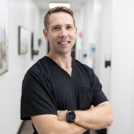 Male dentist in black scrubs smiling in modern dental office hallway.