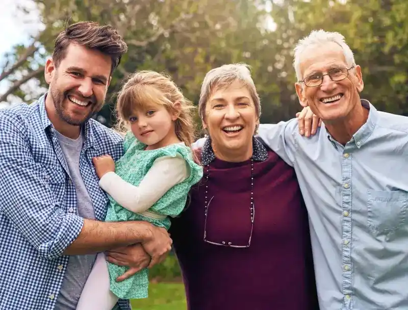 Four-generation family smiling together outdoors in natural sunlight among green trees.